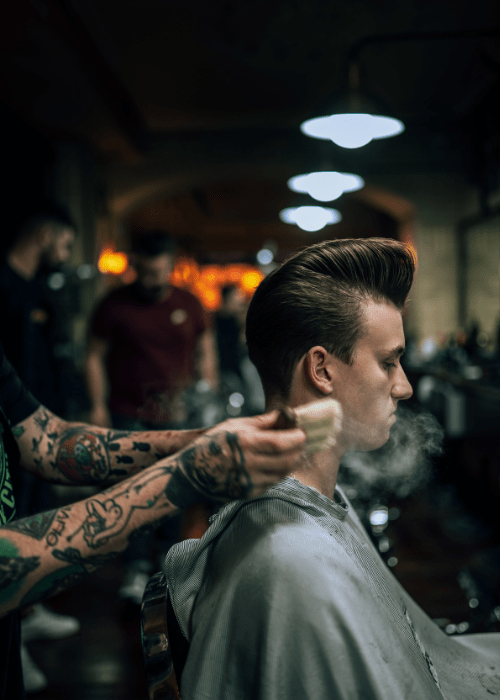 Man getting styled with firm hold pomade by tattooed barber in a dimly lit barbershop